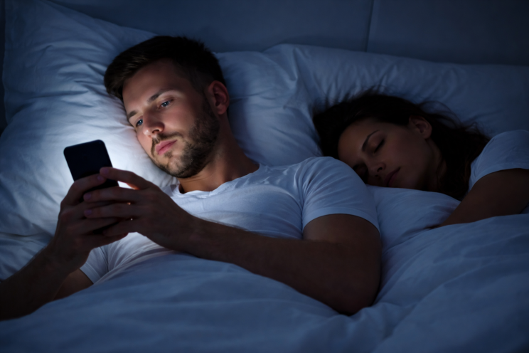 Man lying in bed at night secretly looking at his phone while his partner sleeps beside him in a dimly lit bedroom, suggesting relationship secrecy.