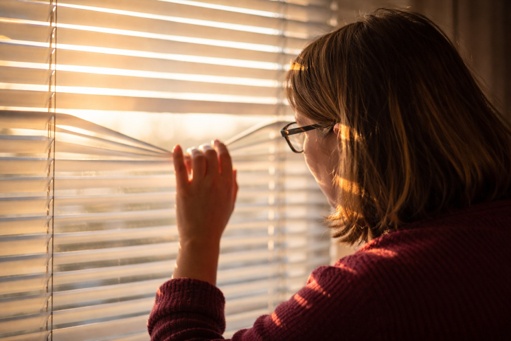 Woman standing by a window looking through blinds as early morning sunlight enters the room, symbolising suspicion and searching for the truth in a relationship.