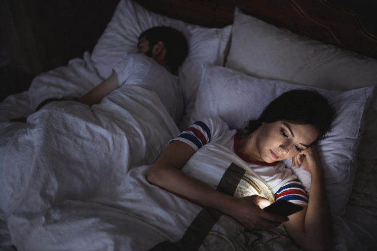 Woman lying awake in bed at night looking at her mobile phone while her partner sleeps beside her, reflecting suspicion and concern in a relationship.
