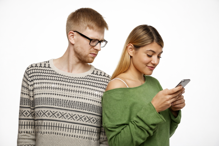 Young man with a suspicious expression glancing at his girlfriend’s phone while she texts on her smartphone.