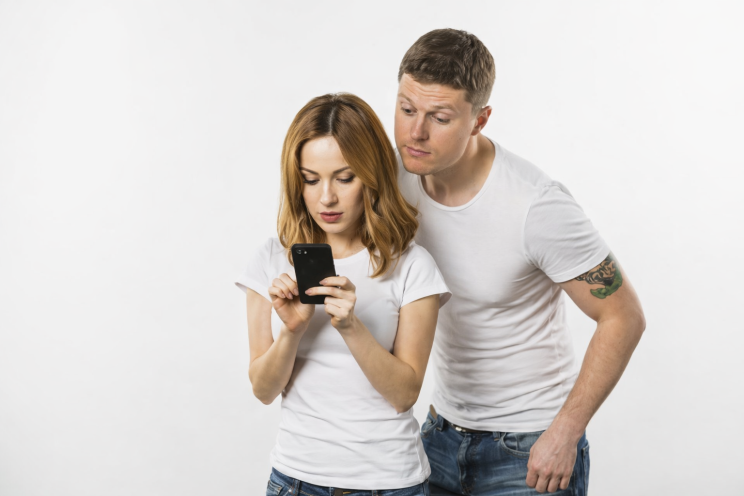 Young man looking over his girlfriend’s shoulder while she reads messages on her smartphone against a white background.