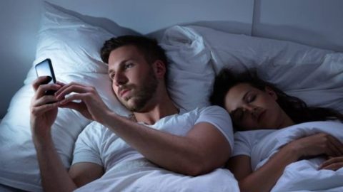 A man discreetly using his phone in the dimly lit bedroom while his unsuspecting wife is sound asleep, symbolizing potential evidence of an affair.