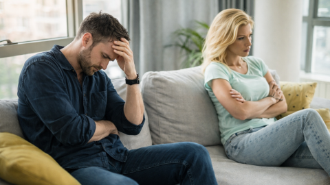 A couple sits apart on a sofa in a modern living room, the man holding his head in frustration while the woman sits with arms crossed, both appearing deep in thought.