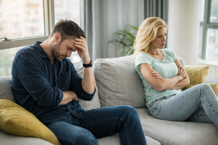 A couple sits apart on a sofa in a modern living room, the man holding his head in frustration while the woman sits with arms crossed, both appearing deep in thought.
