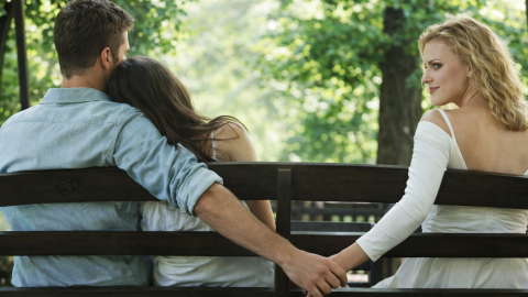 Man sitting on a bench with a woman leaning on his shoulder while secretly holding hands with another woman behind the bench, suggesting hidden relationship tension