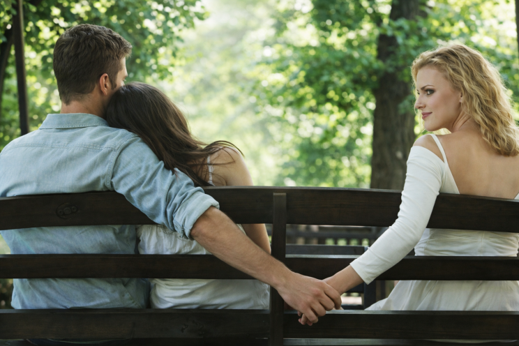 Man sitting on a bench with a woman leaning on his shoulder while secretly holding hands with another woman behind the bench, suggesting hidden relationship tension