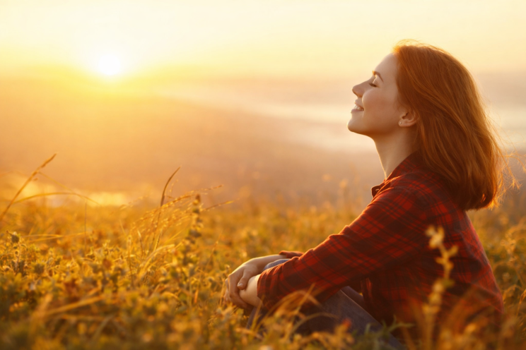 Woman sitting peacefully in a golden field at sunset, eyes closed and smiling, reflecting emotional healing and personal growth after relationship challenges