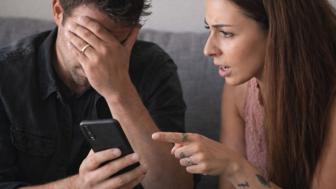A couple sitting closely on a couch looking at a smartphone, the man covering his face in stress while the woman points at the screen during a tense conversation