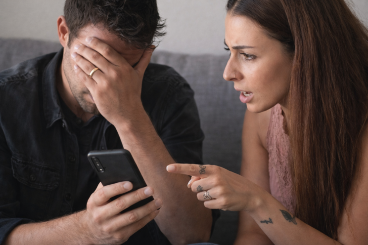 A couple sitting closely on a couch looking at a smartphone, the man covering his face in stress while the woman points at the screen during a tense conversation