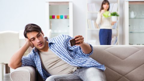 Man looking at his phone on the couch while a woman watches from behind