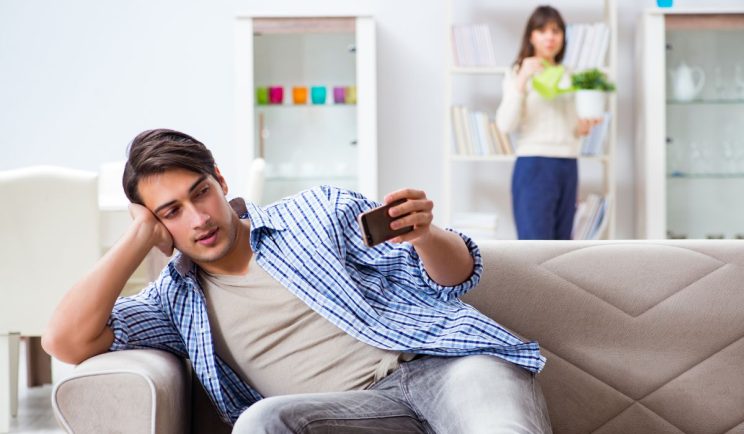 Man looking at his phone on the couch while a woman watches from behind