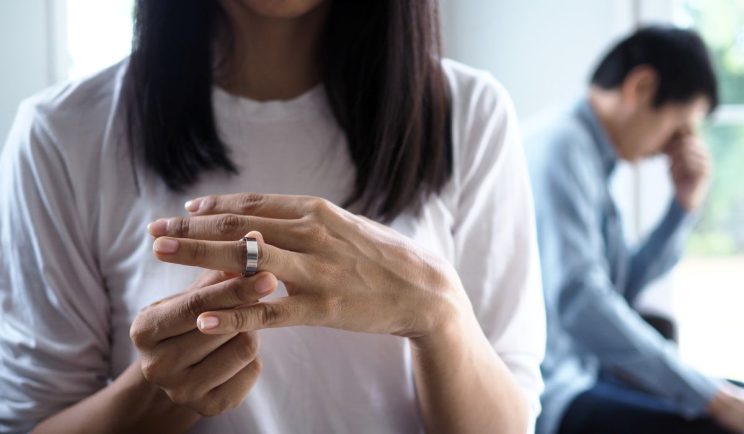 A woman taking off her wedding ring with a distressed man sitting in the background