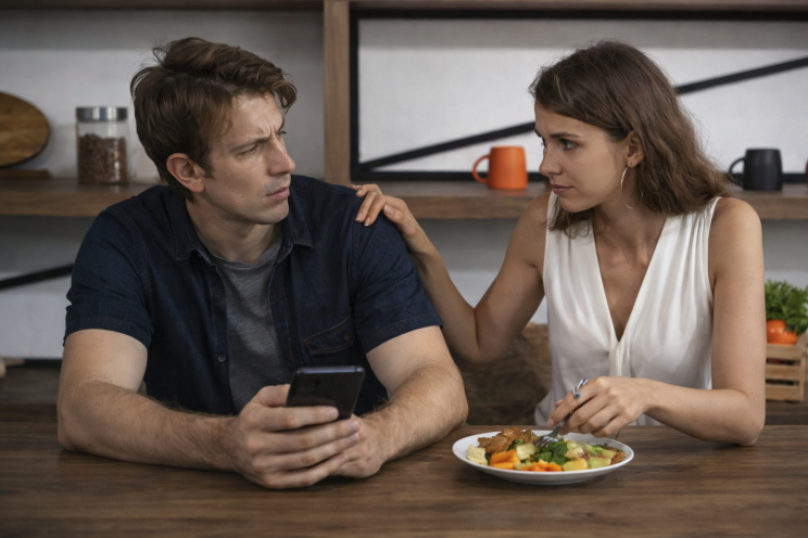 Man holding a phone at a dining table while his partner touches his shoulder, showing relationship tension and concern.