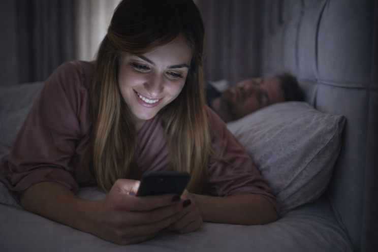 Woman lying in bed smiling at her phone while her partner sleeps beside her in the background.